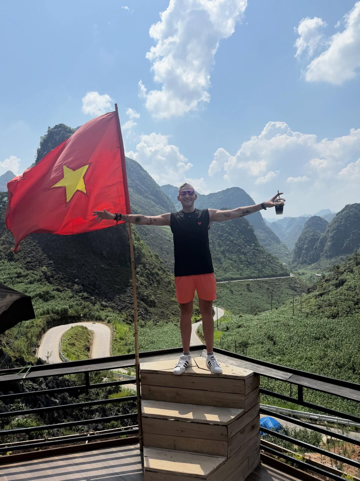 Foreign-tourist-holding-a-coffee-next-to-the-Vietnam-flag-at-the-spectacular-Ma-Pi-Leng-Pass-in-Ha-Giang