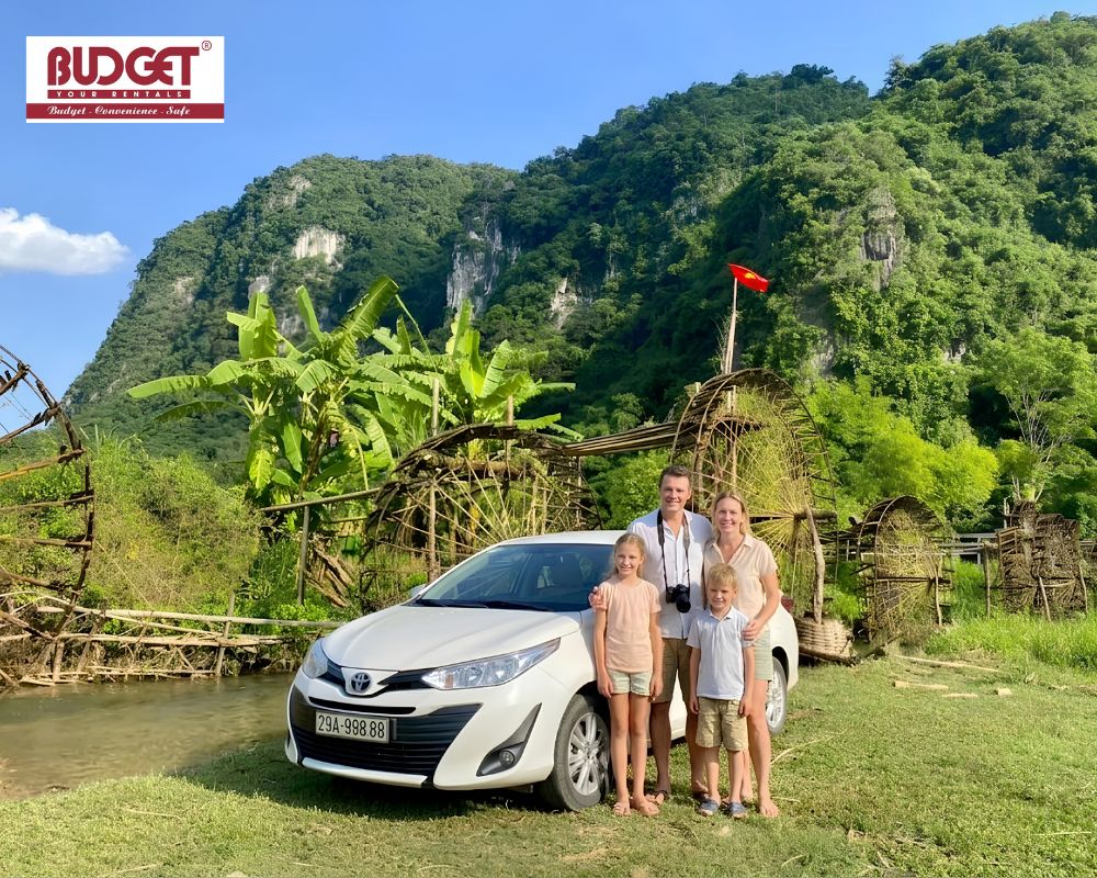 Happy-family-posing-with-a-white-Toyota-sedan-by-traditional-bamboo-water-wheels-and-karst-mountains-in-Pu-Luong