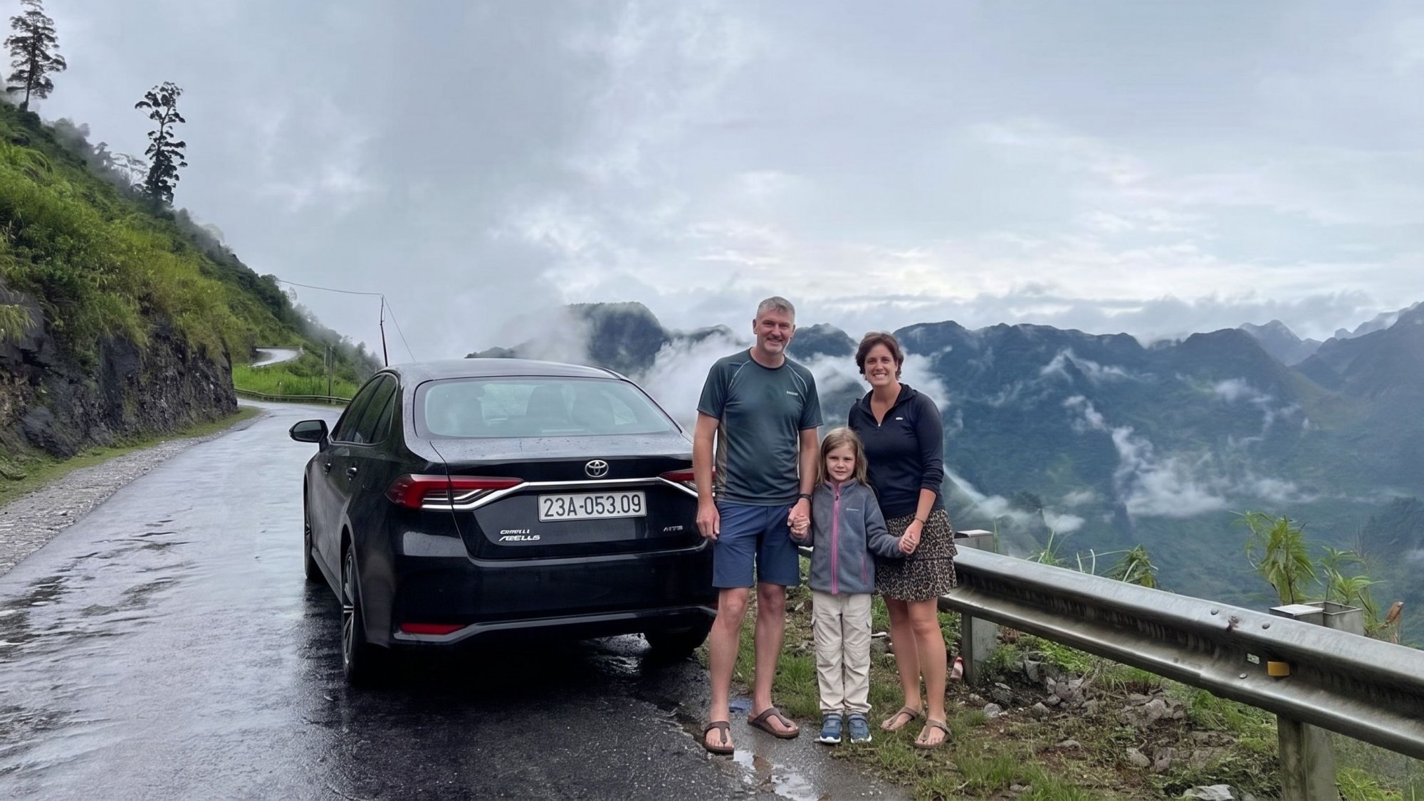 Tourist-family-of-three-posing-next-to-their-4-seater-rental-car-on-a-cloudy-mountain-pass-Ha-Giang