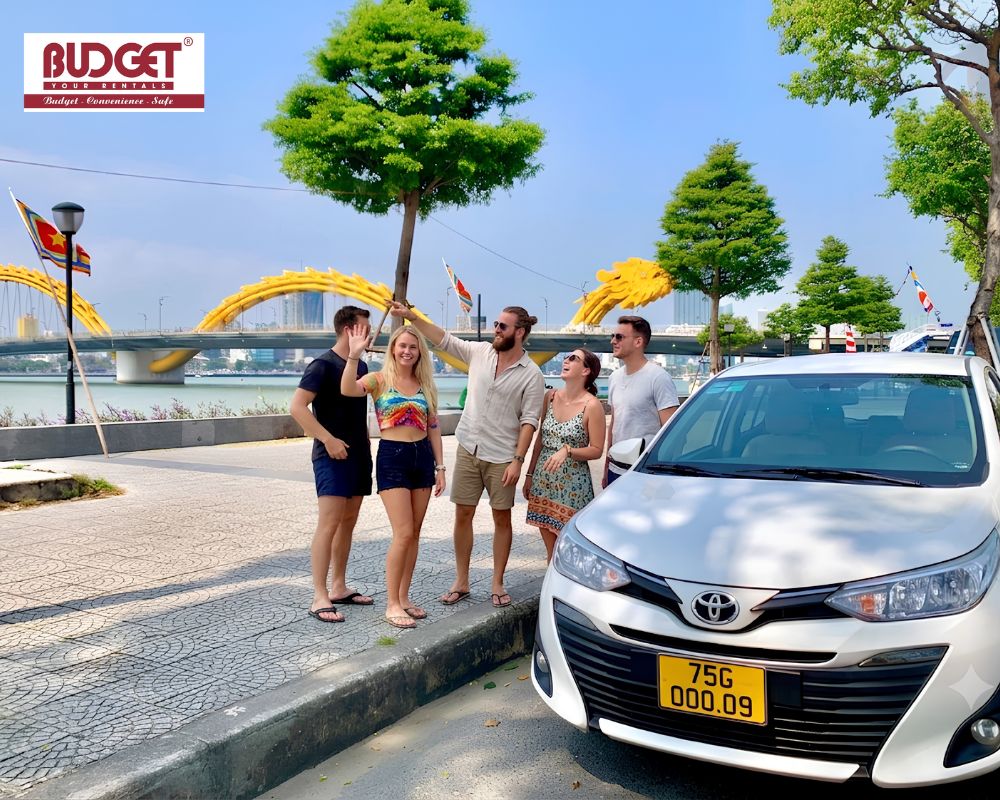 Group-of-young-tourists-posing-beside-a-white-Toyota-sedan-with-the-famous-yellow-Dragon-Bridge-in-Danang