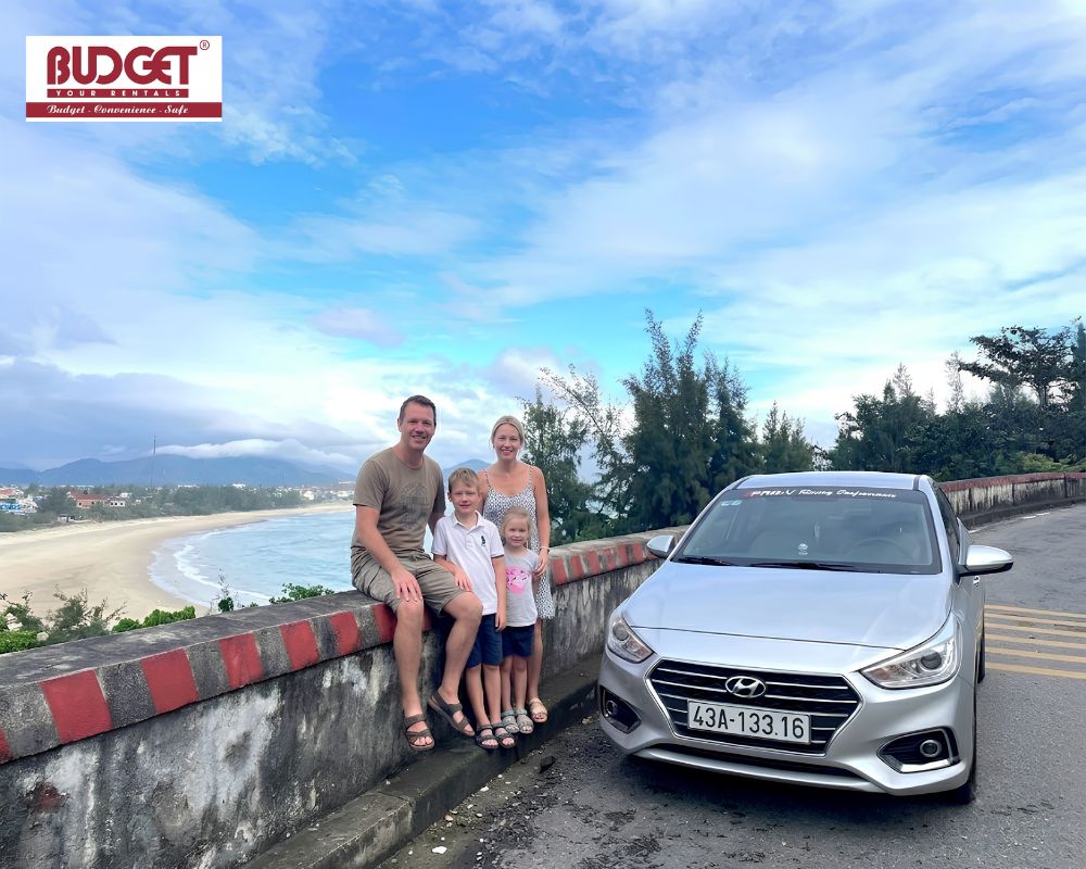Happy-family-tourists-posing-next-to-a-silver-Hyundai-sedan-on-a-scenic-coastal-road-at-Hai-Van-Pass